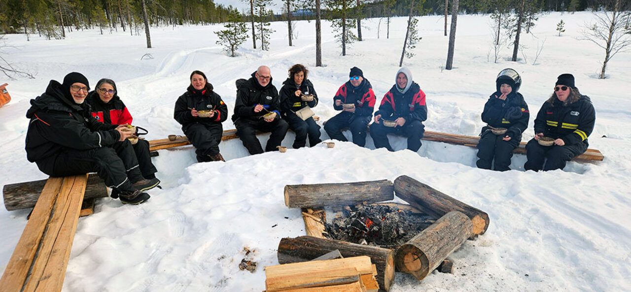 The photograph shows nine people sitting in a half circle on wooden benches around a fireplace. The ground is covered with snow. All persons hold a bowl and spoons and look directly into the camera.