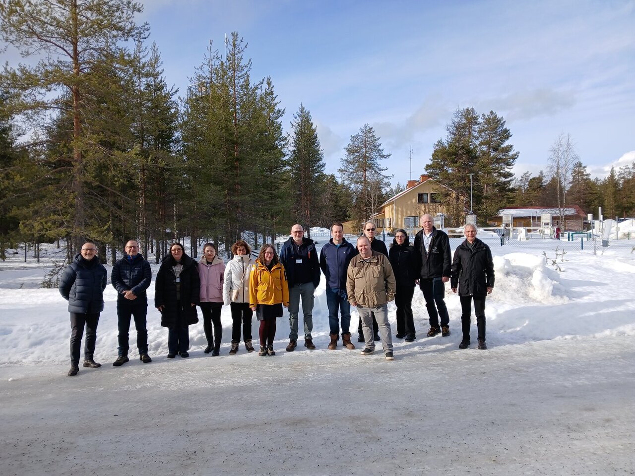 The photograph shows 13 people standing in front of large trees in a snowy environment. The persons' sights are directed to the camera.